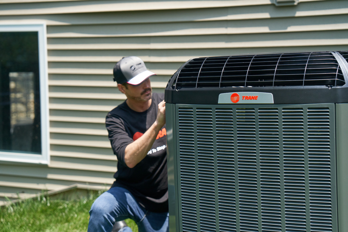 Two HVAC technicians installing or servicing a large Trane outdoor air conditioning unit next to a white brick home; one technician kneels while handling components, and the other steadies the unit.